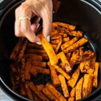 A hand holding one sweet potato fries over the cooked fries in the air fryer showing how crisp it is