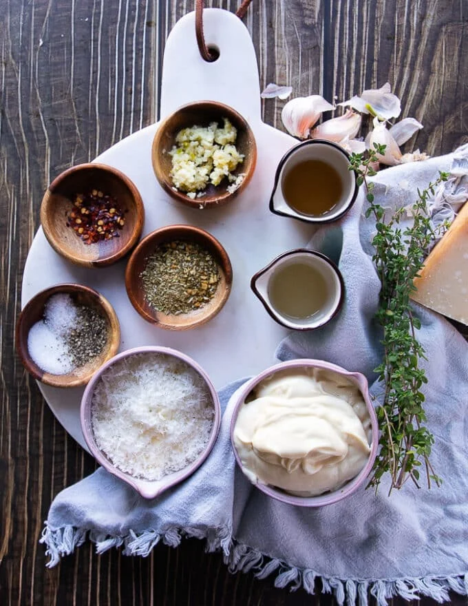 Ingredients for the garlic parmesan sauce in a board. A bowl of mayo, some crushed garlic in a bowl, lemon juice in a bowl, some italian seasoning in a bowl, some salt and pepper in a bowl, some apple cider vinegar and a bowl of grated parmesan chese