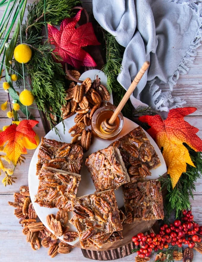 The pecan pie bars all arranged on a white marble surrounded by extra whole pecans and a small honey saucepan on the side