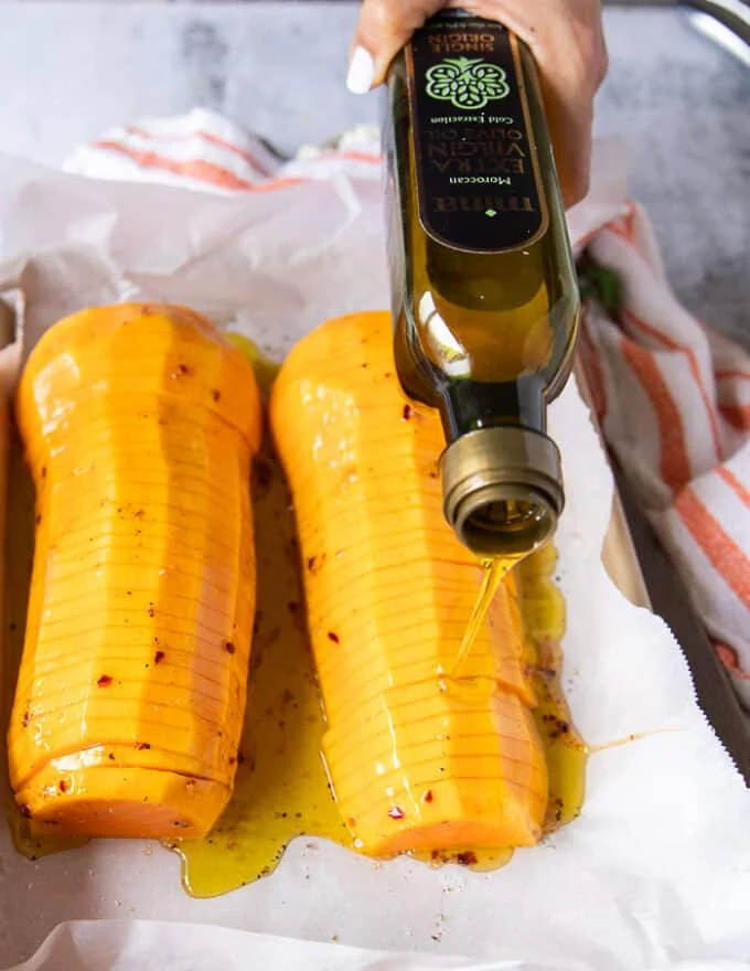 A hand pouring olive oil over seasoned butternut squash halves on a parchment lined baking sheet