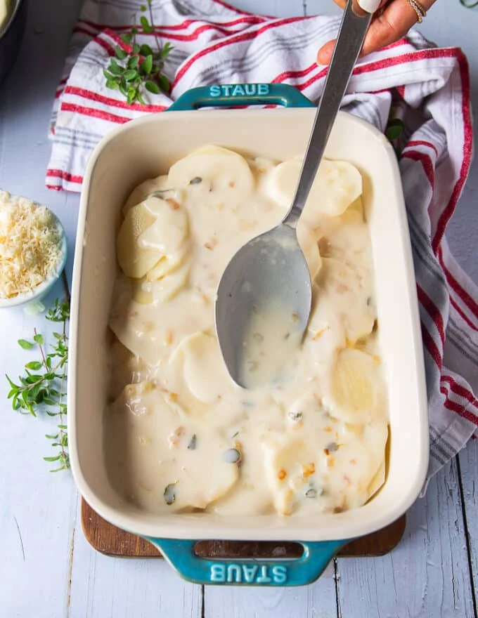 A ladle pouring half of the cream sauce over the potatoes layering to make the scalloped potatoes