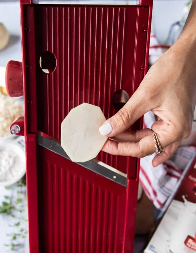 A hand holding a thinly sliced potato in front of a mandolin slicer