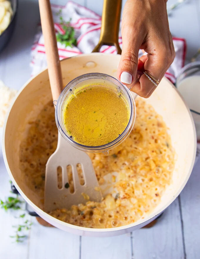 A hand pouring the stock in to the onions and flour mixture to start making the sauce