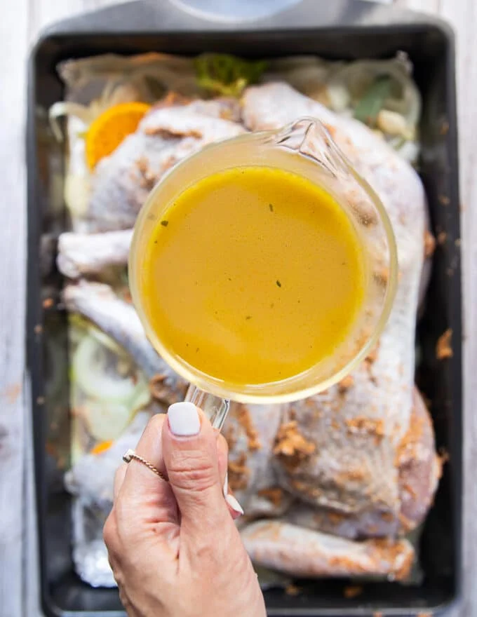 The buttered turkey placed over the onions and oranges and veggies on the roasting pan and a hand ready to pour some stock over it.