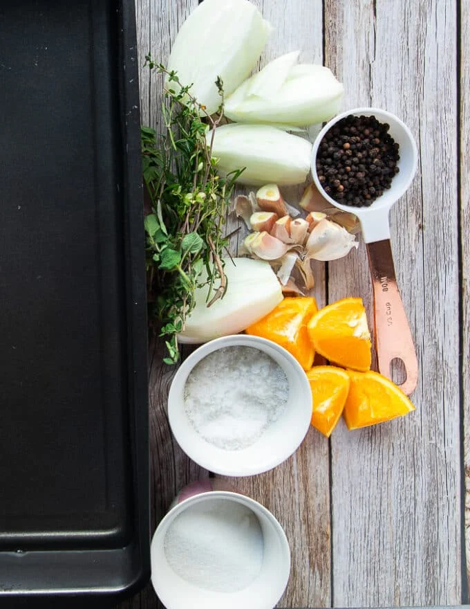 turkey brine ingredients on a table including a large roasting pan with water, a bowl of salt, bowl of sugar, orange slices, quartered onions, whole garlic cloves, fresh herbs, whole peppercorns in a bowl