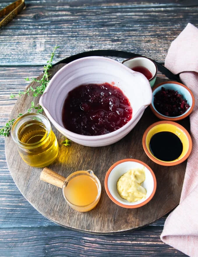 Ingredients for sweet potato salad dressing in a wooden board including a bowl of cranberry sauce, a bowl of mustard, some vinegar in a bowl, some balsamic vinegar in a bowl, some olive oil, and dried cranberries in a bowl