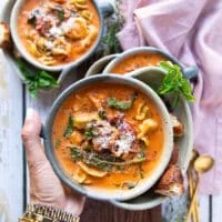 A hand holding one bowl of tortellini soup close up showing the cooked tortellini in a creamy tomato broth with basil leaves and garnished with fresh parmesan cheese