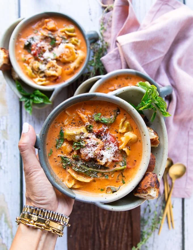A hand holding one bowl of tortellini soup close up showing the cooked tortellini in a creamy tomato broth with basil leaves and garnished with fresh parmesan cheese