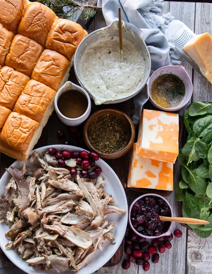 All ingredients for the turkey sliders recipe on a cutting board. A pack of Hawaiian dinner rolls, some sliced marbled cheese, some leftover turkey meat, a bowl of cranberry sauce, a bowl of gravy, some fresh spinach, garlic parmesan sauce, and garlic butter for brushing