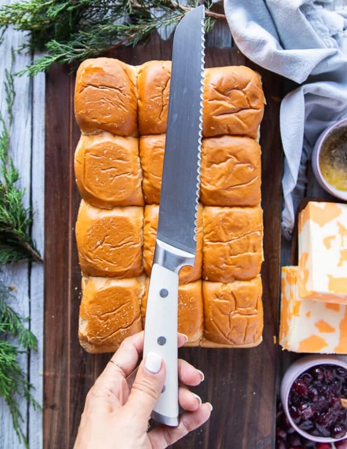 A hand holding a serrated knife to cut cross sectionally the hawaiian rolls bread or slider bread.