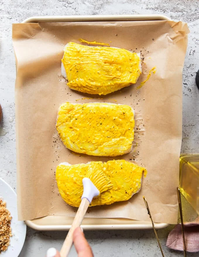 a hand brushing some spicy mustard over the tops only of the cod fish