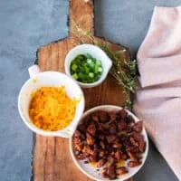 A board with small bowls of topping for the baked soup recipe including the crumbled bacon in a plate, some shredded cheese in a bowl and some green onions minced