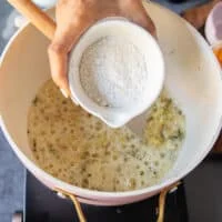 A hand adding in the flour into the onion and butter soup base in the pan