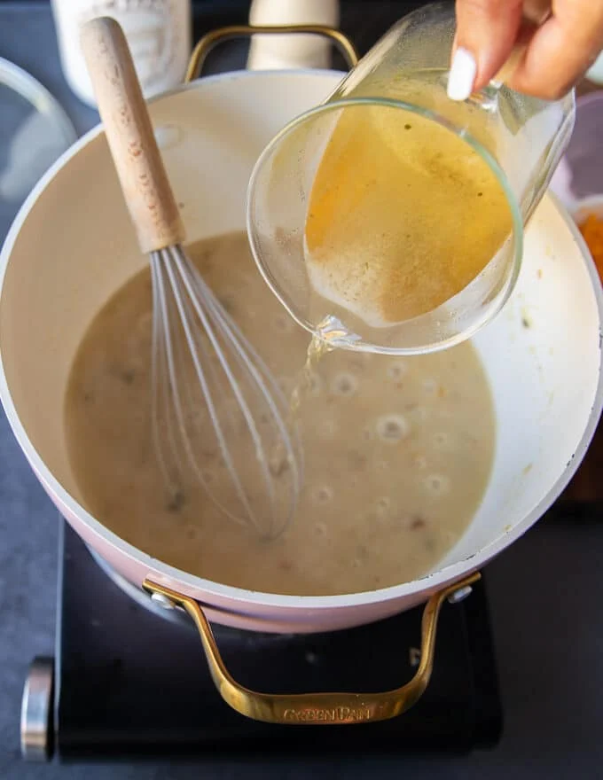 A stock is added gradually to the flour mixture and a hand holding a whisk is whisking it in gradually