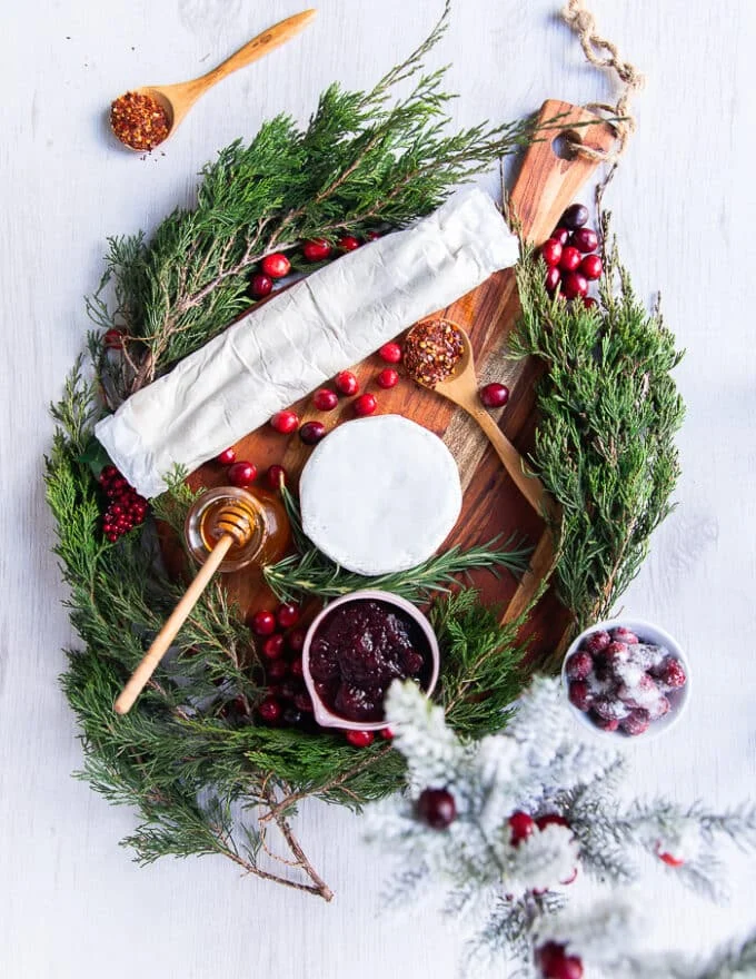 Ingredients needed to make cranberry brie bites on a wooden board including a block of bire cheese, some cranberry sauce and a roll of puff pastry. Some hot honey extra on the side along with fresh rosemary sprigs