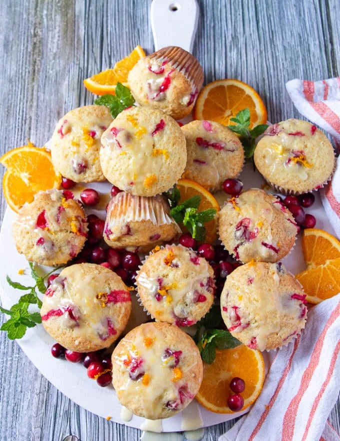 A final board of orange cranberry muffins glazed and ready to serve surrounded by an orange tea towel and some orange slices, some mint leaves and some fresh cranberries