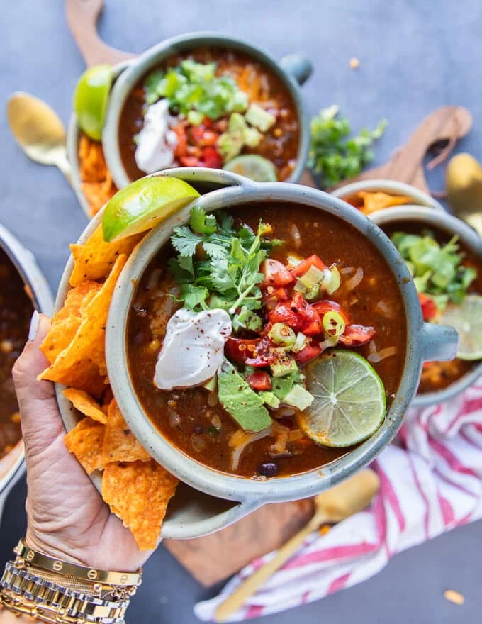 A. hand holding one bowl of taco soup showing a close up of the beans, tomato broth, corn, minced beef and the toppings like avocados, sour cream, cheese, a lime slice and cilantro