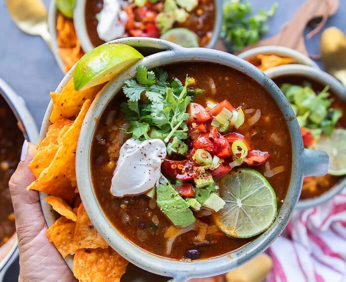 A. hand holding one bowl of taco soup showing a close up of the beans, tomato broth, corn, minced beef and the toppings like avocados, sour cream, cheese, a lime slice and cilantro