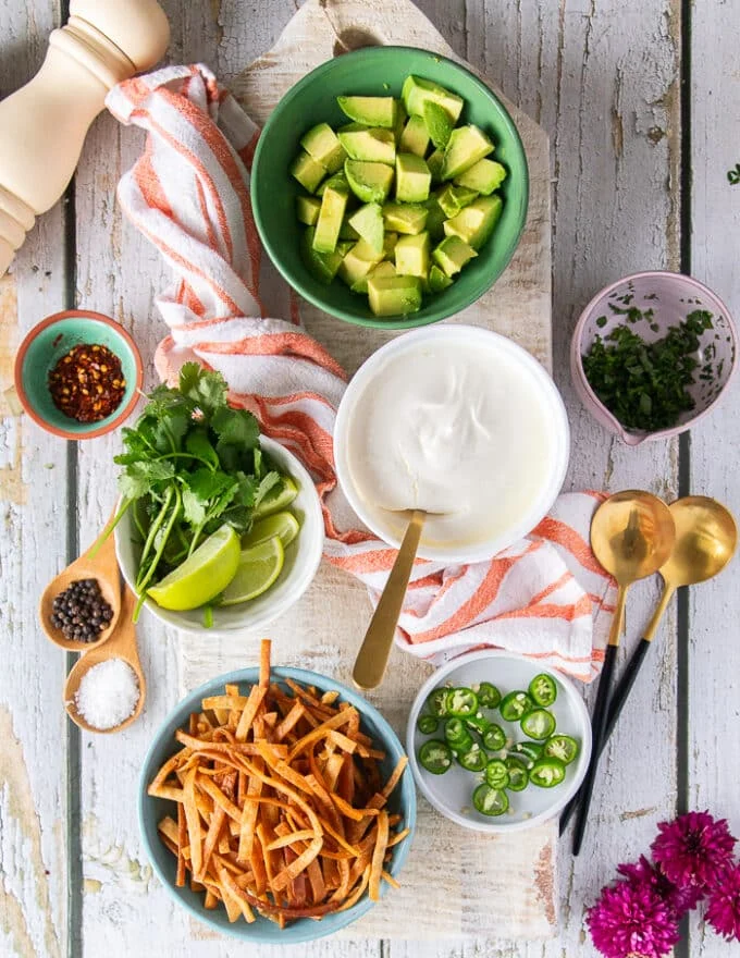 Small bowls of toppings to be added over the bowls of soup. There's a bowl of sour cream, a bowl of diced avocados, a bowl of the tortilla strips, a bowl of lime and cilantro leaves, a bowl of green onions, a bowl of jalapenos