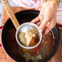 A hand holding the plate of different spices like the oregano, garlic powder, cumin, chilli powder, salt and pepper and adding it into the pot of soup