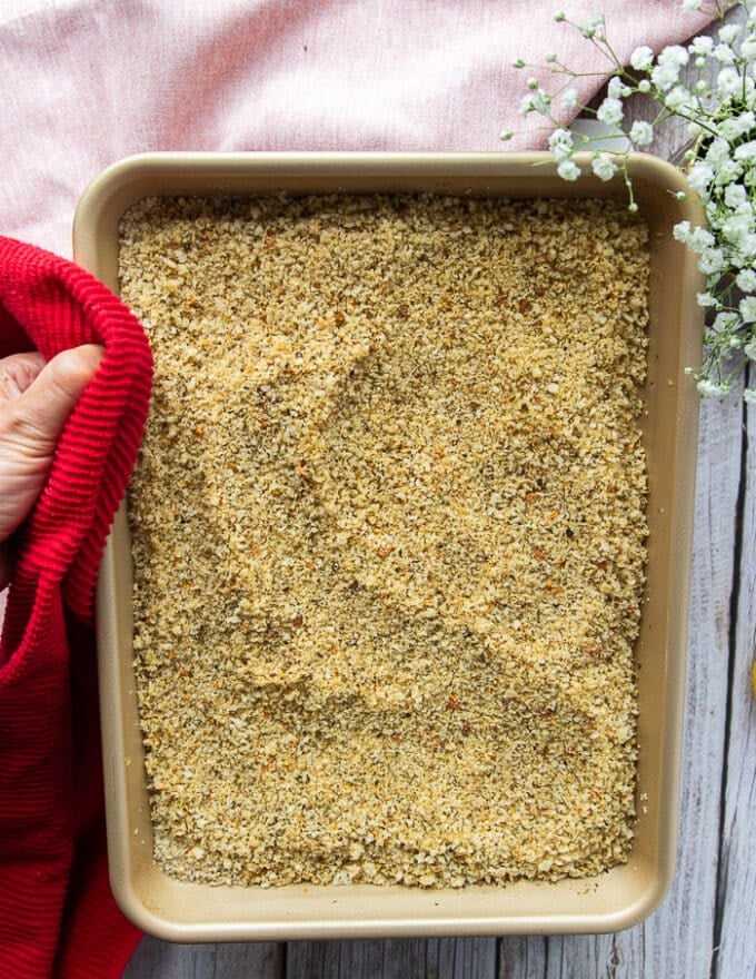 A hand holding a sheet pan of the toasted panko bread crumbs mixture