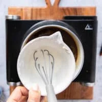A hand adding in a bowl of cornstarch slurry which is cornstarch and water mixed in a cup