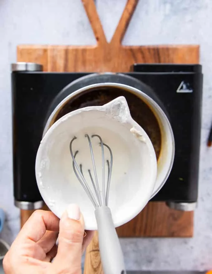 A hand adding in a bowl of cornstarch slurry which is cornstarch and water mixed in a cup