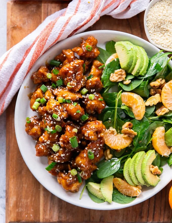 close up of a serving of homemade orange chicken on a plate showing the sticky orange chicken perfectly cooked and garnished with sesame and green onions along side some avocado salad