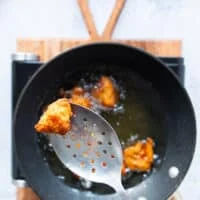 chicken frying in the oil has turned golden and is ready to be removed from the oil. A hand holding a spoon to remove the chicken