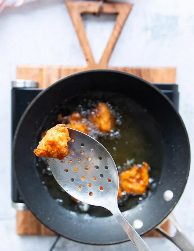 chicken frying in the oil has turned golden and is ready to be removed from the oil. A hand holding a spoon to remove the chicken
