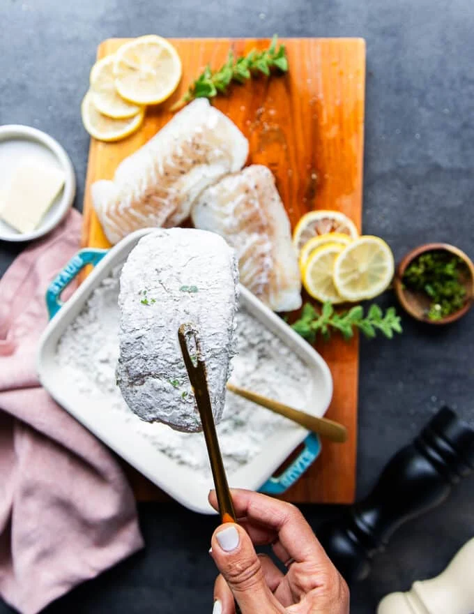A hand holding a cod fish dusted lightly with flour and showing how lightly coated it is above the flour plate