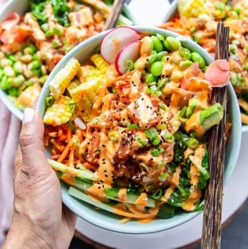 A hand holding a poke bowl very close with chopsticks. The poke bowl includes sushi tuna, rice, carrots, edamame , seaweed salad, pickled ginger, cucumbers all drizzled with spicy mayo