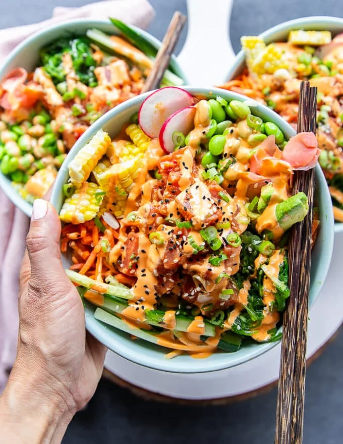 A hand holding a poke bowl very close with chopsticks. The poke bowl includes sushi tuna, rice, carrots, edamame , seaweed salad, pickled ginger, cucumbers all drizzled with spicy mayo