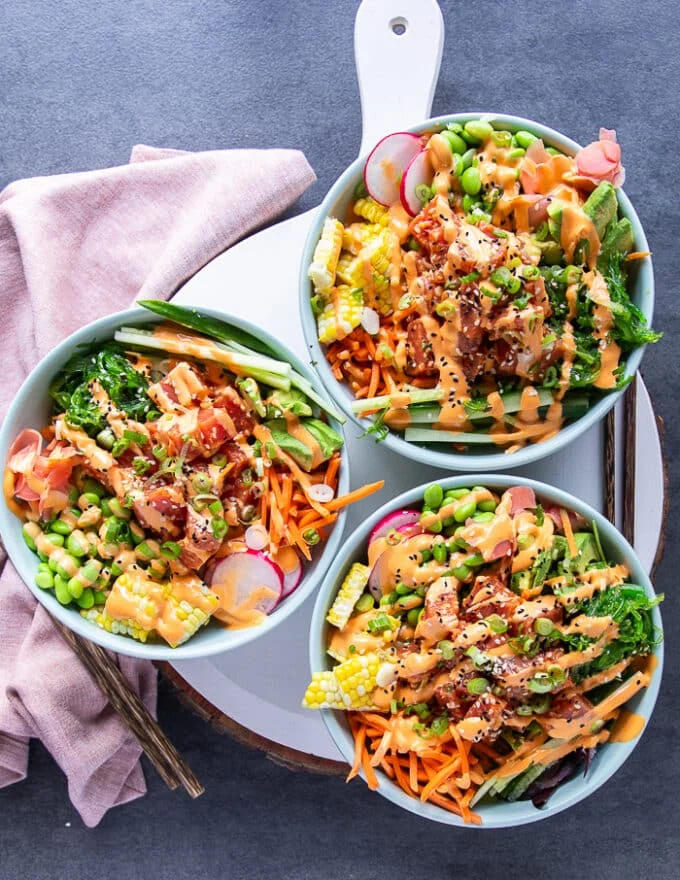 three poke bowls assembled and ready on a board. Drizzled with spicy mayo, topped with sesame seeds