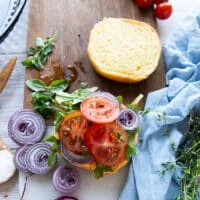 The toppings are loaded over the burger bun including some greens, sliced onions, sliced tomatoes