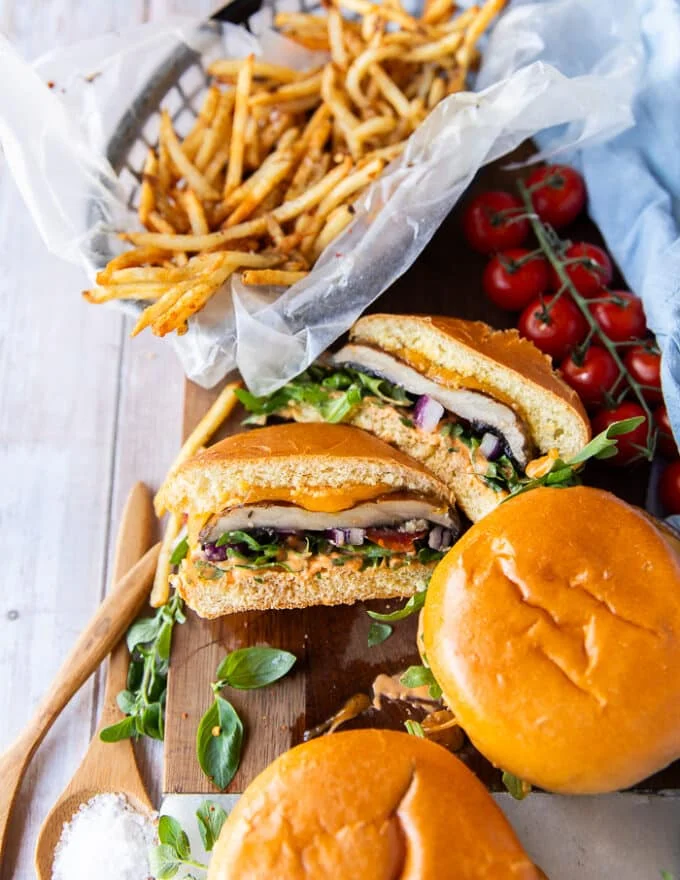 Three portobello mushroom burgers on a wooden plate with one of them cut in half showing the texture and filling of the burger and the portobello mushroom. The burgers are surrounded by a plate of french fries