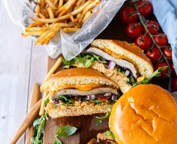 Three portobello mushroom burgers on a wooden plate with one of them cut in half showing the texture and filling of the burger and the portobello mushroom. The burgers are surrounded by a plate of french fries