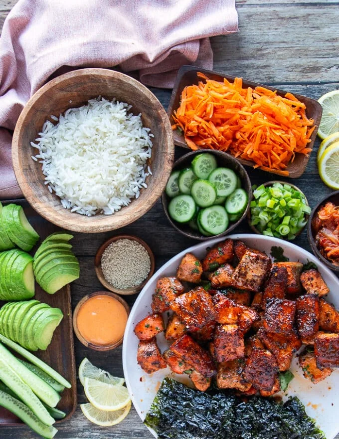 ingredients needed to make salmon bowl: cooked salmon bites in a bowl, coked rice in a bowl, shredded carrot on a plate, sliced avocados on a board, sliced cucumbers and green onions on a board, some spicy mayo, sesame seeds in a bowl, shredded seaweed on a bowl