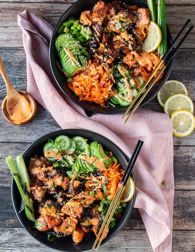 two salmon bowls on a table with salmon, rice, avocados, cucumber, pickled ginger, seaweed and spicy mayo drizzle