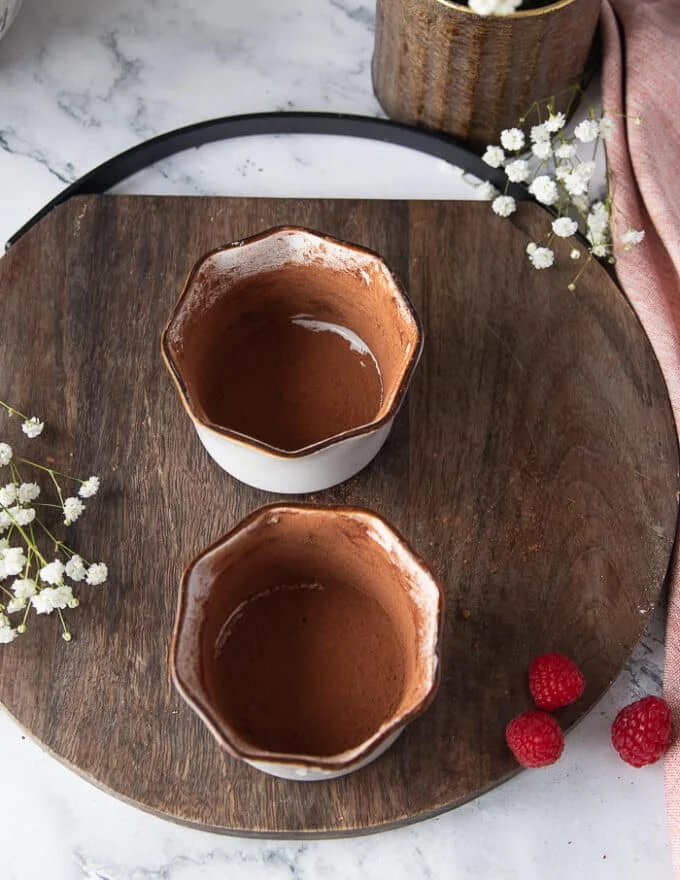Two ramekins perfectly coated in butter and cocoa powder on a wooden tray