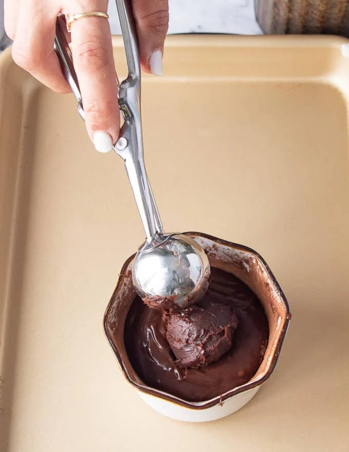 A hand holding an ice cream scoop of the truffle mixture and scooping in in the center of the cake batter in the ramekin before baking