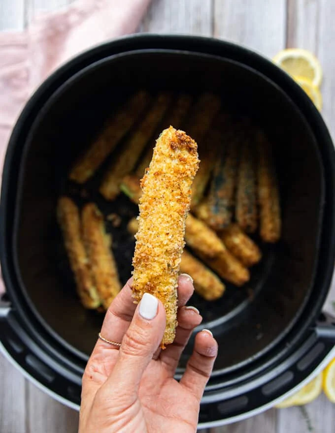 A hand holding one zucchini fries right out of the air fryer showing the texture and color of it