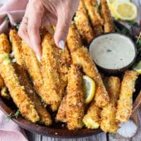 A hand holding one crunchy zucchini on a plate of zucchini fries surrounded by parmesan dip