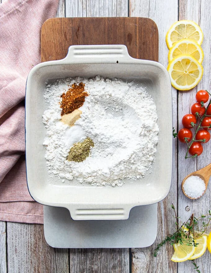 Flour coating ingredients on a board including a plate of flour, some garlic powder, paprika, salt, pepper, and oregano in separate bowls