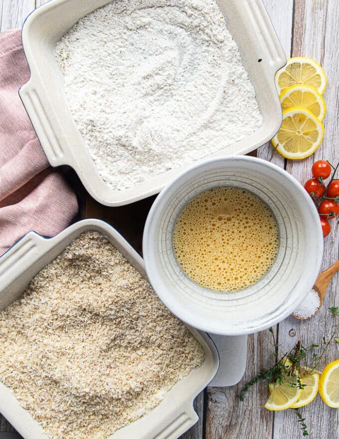 The breading station ready on a wooden board including the flour coating with the spices mixed in in a plate, the eggs beaten in a bowl and the panko coating with the spices and parmesan cheese mixed in a shallow plate