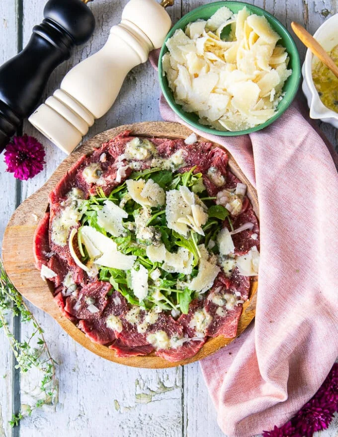 A plate of beef carpaccio dressed up and ready with a caper vinegar mustard vinaigrette and topped with some parmesan cheese and arugula in the middle