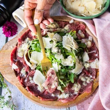 A hand holding a fork with a beef carpaccio close up showing the thinly sliced meat, the light vinaigrette and topping of diced onions, arugula and parmesan shavings