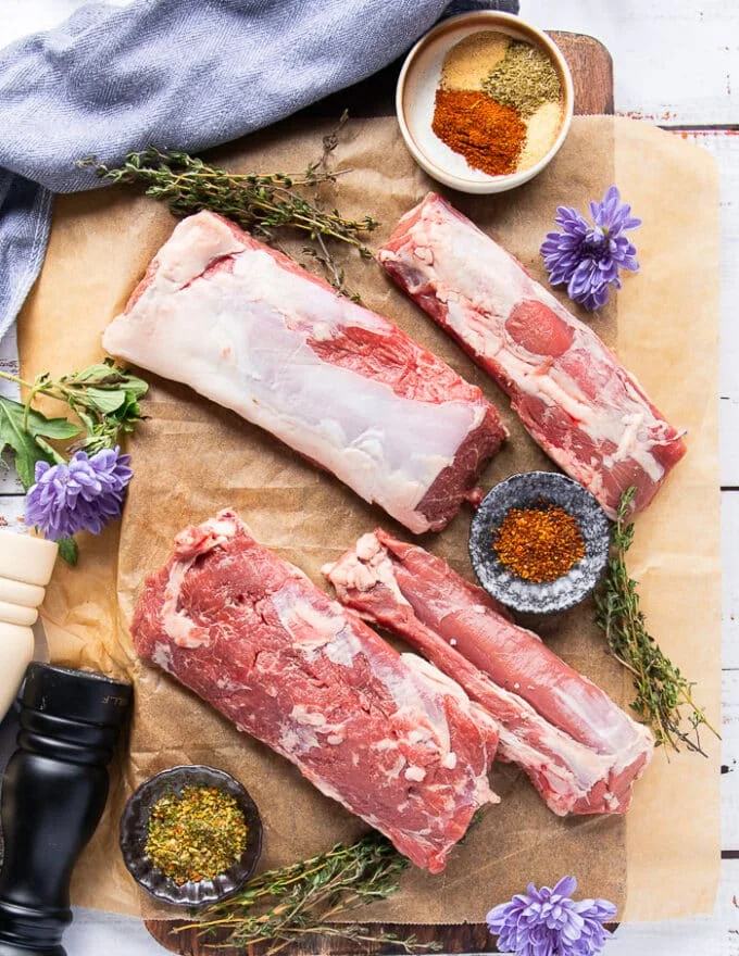 A set of lamb loin deboned and lamb sirloin and lamb tenderloin deboned on a parchment paper surrounded by a small bowl of chili flakes and some fresh thyme leaves