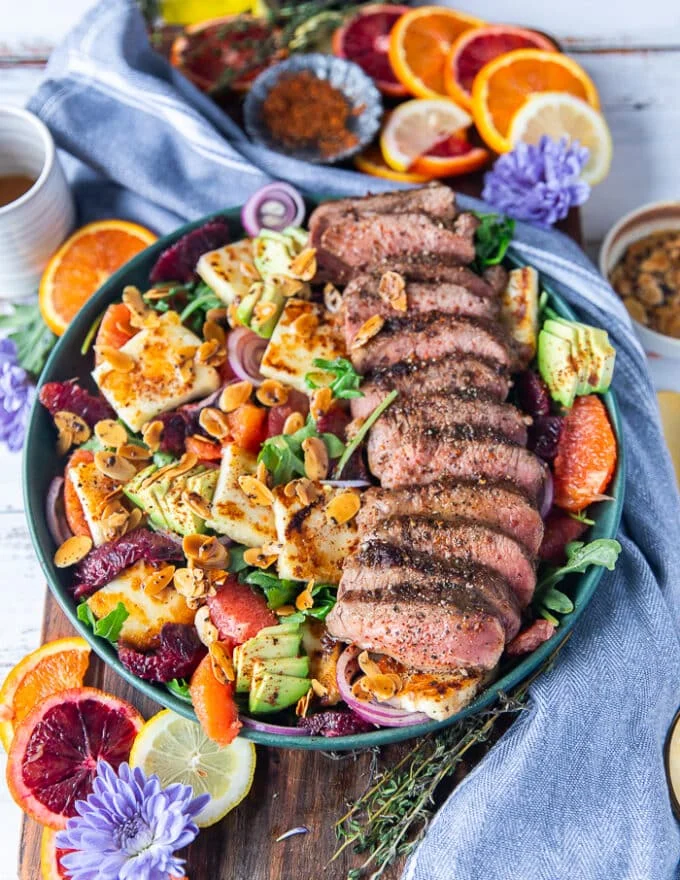 A vibrant plate of lamb loin roasted and sliced served over a halloumi salad with orange segments, arugula, onions, avocados and some crunchy almonds