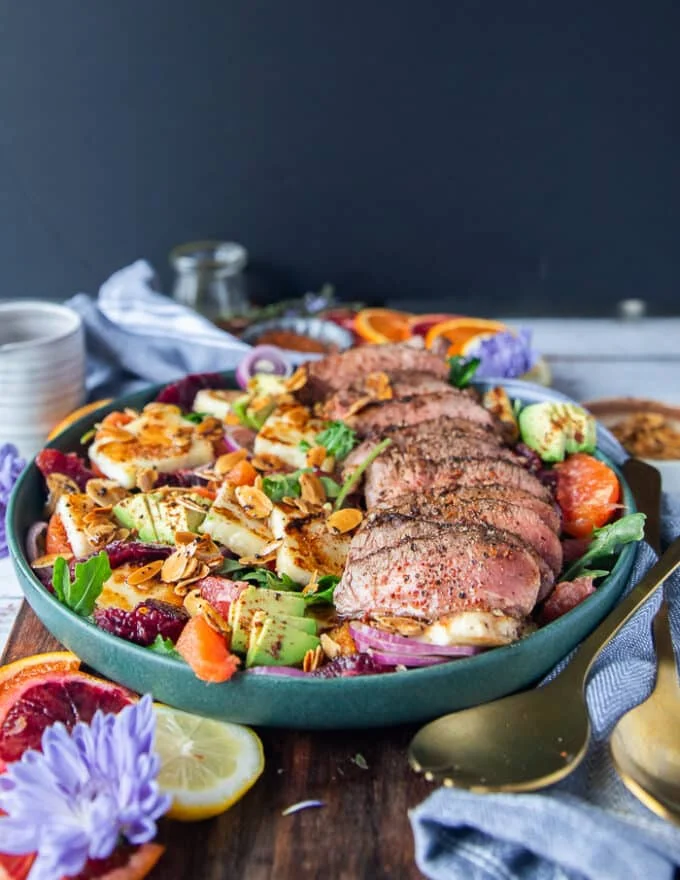 A close up plate of the sliced lamb loin and lamb sirloin over a bed of halloumi salad and citrus segments , avocados, almonds, onions and a citrus honey dressing